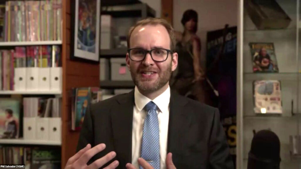 A man in glasses and a suit sitting in a library and talking to the camera. He is gesturing with his hands.