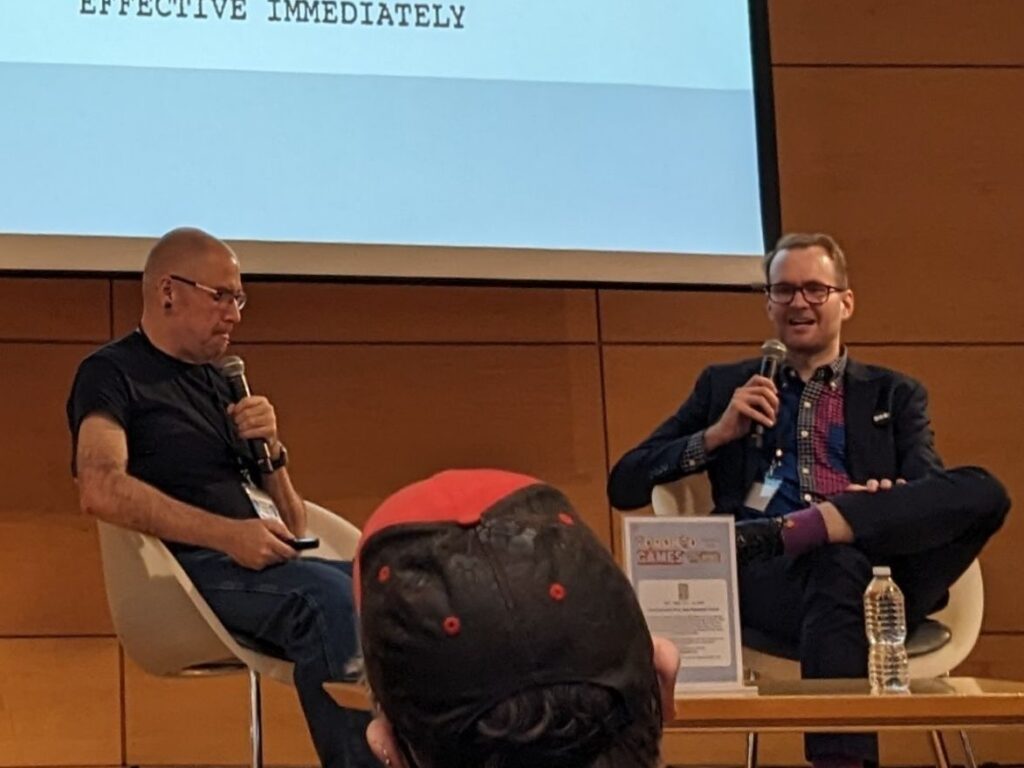 Two men sitting on a stage, talking into microphones.