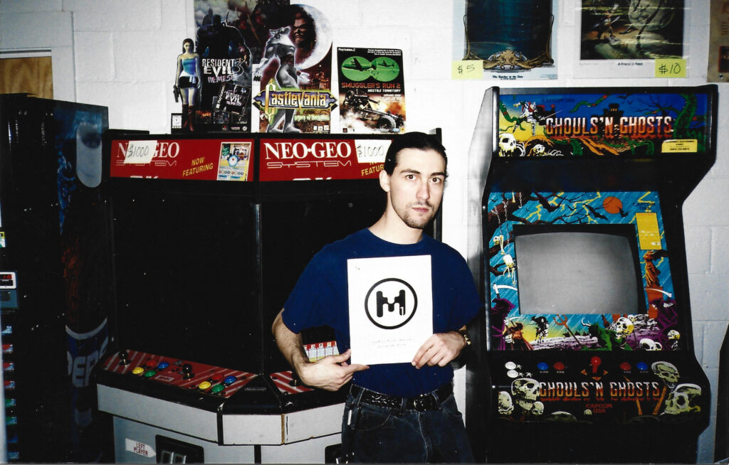 MAGFest founder Joseph Yamine at his game store, Captain Gamestation, holding a program for MAGFest 1. He is surrounded by various arcade games and video game memorabilia.