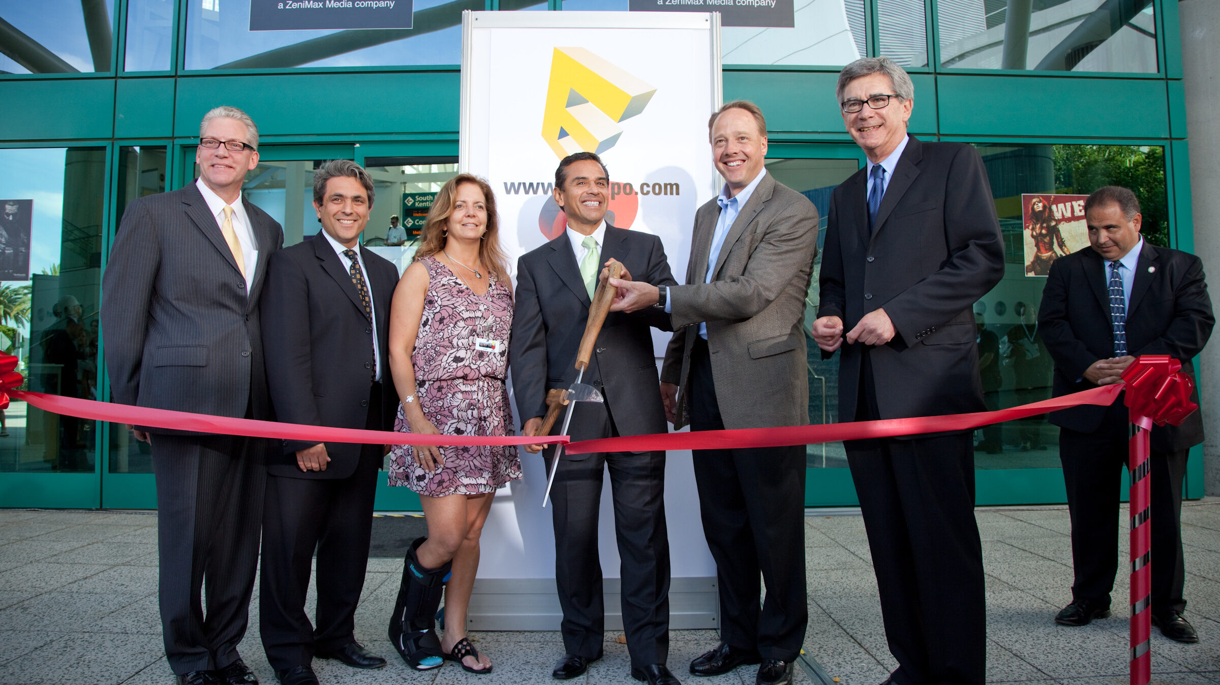 A group of executives cutting a ribbon in front of the Los Angeles Convention Center. Mary Dolaher, a woman in a pink dress, is wearing a leg brace.
