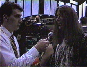 Jeff Minter, a man with long hair, glasses, and a cardigan, talks to a reporter in front of an Atari Jaguar kiosk.