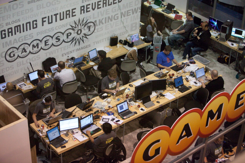A distant top-down shot of GameSpot's press booth at E3. Journalists are crammed into a small space writing articles.