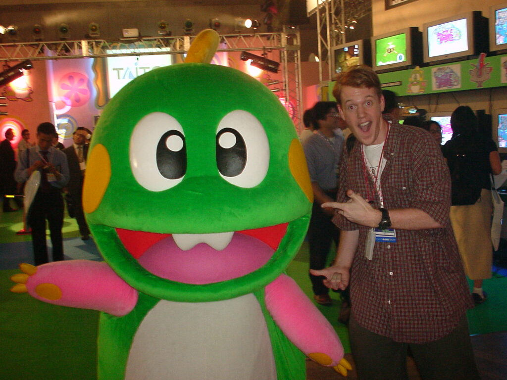 Colin Williamson with a mascot character of Bub from Bubble Bobble. Super Puzzle Bobble is being played at Taito's booth in the background.
