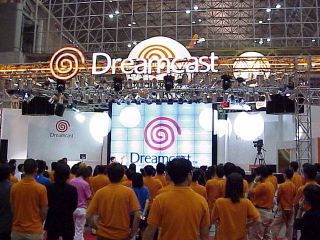 A huge pavilion stage promoting the Dreamcast at Tokyo Game Show 1999 Autumn. Most people in the audience are wearing orange polo shirts, the same color as the Dreamcast logo.