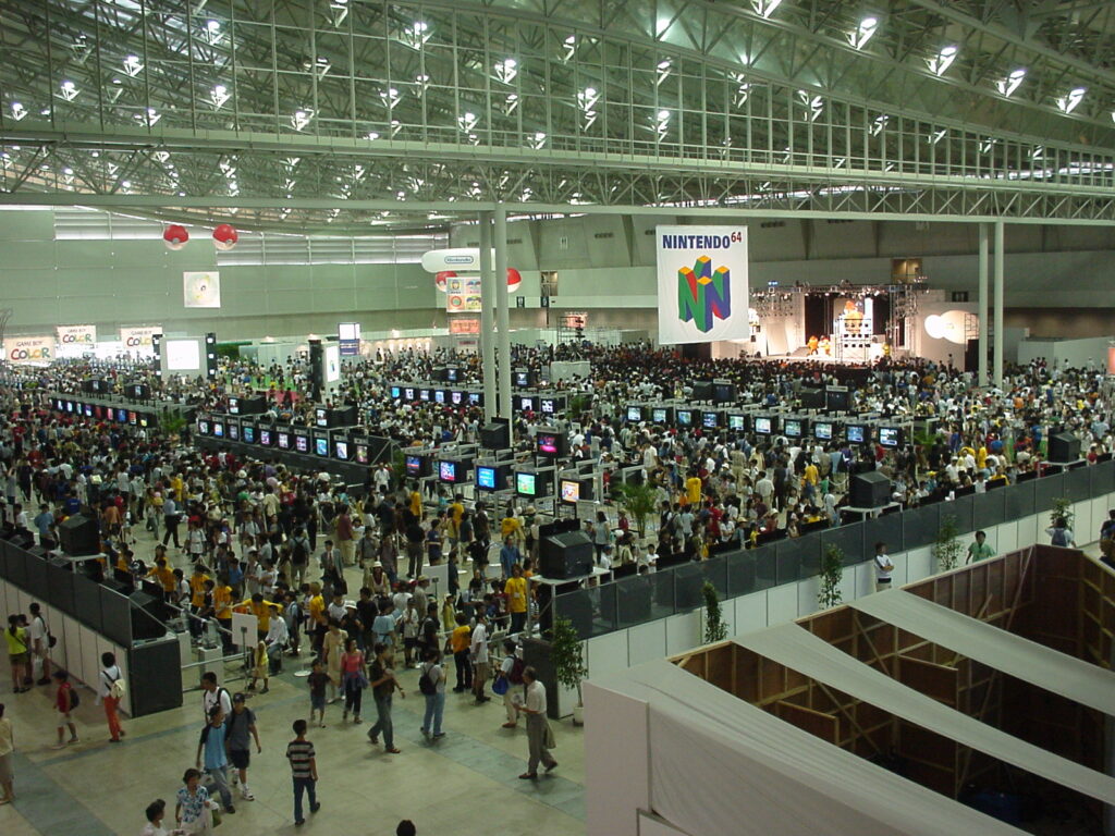 A view of Nintendo Space World 2000. Banners for the Nintendo 64 and Game Boy Color are prominently hung from the ceiling. Pokeballs are also suspended from the rafters in the distance. There is also a stage, where performers appear to be dressed as Donkey Kong and Diddy Kong.