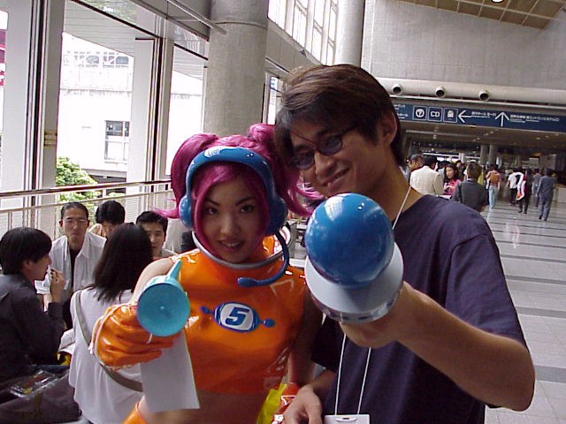 Space Channel 5 producer Tetsuya Mizuguchi posing with a person dressed as Ulala, the protagonist of Space Channel 5. Mizuguchi is holding Ulala's microphone to the camera.